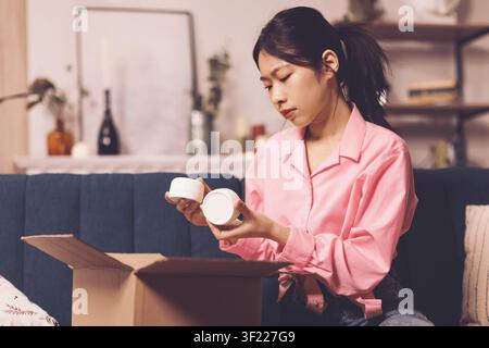 Junge asiatische Frau, die Hautpflegeprodukte mit konzentriertem Gesichtsausdruck zu Hause auspackt, Schönheitskonzept Stockfoto