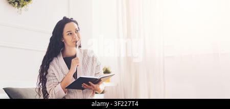 Nachdenkliche junge Frau schreibt in einem Notizbuch zu Hause, gemütliche Inneneinrichtung, konzentrierter Ausdruck Stockfoto