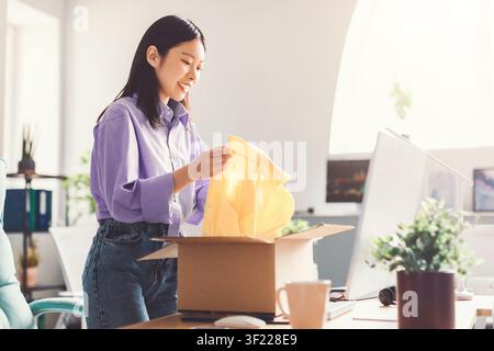 Zufriedene asiatische Frau, die eine Lieferbox in einem hellen modernen Büro auspackt, Begeisterungskonzept Stockfoto