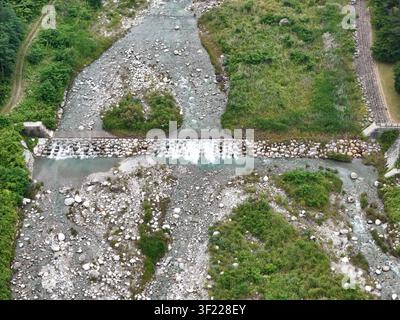 Robustes Flussbett fließt durch felsiges Gelände. Grün säumt das Wasserlauf sanft. Ein kleiner Damm überspannt den Lauf des Baches. Klares Wasser stürzt über Steine Stockfoto