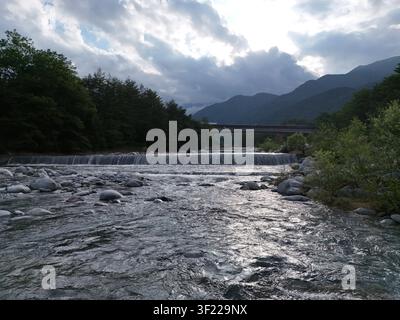 Ein rauschender Fluss fließt über glatte Steine. Ferne Hügel stehen unter einem bewölkten Himmel. Das Flussufer ist gesäumt von üppigen grünen Bäumen. Natürliche Ruhe spiegelt Erbsen Stockfoto