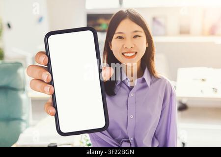 Lächelnde asiatische Frau hält Smartphone mit leerem Bildschirm im Büro, Geschäftskonzept Stockfoto