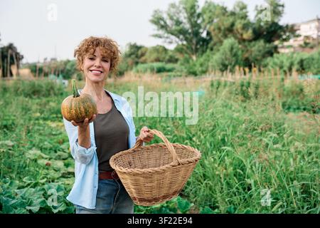 Frau lächelt, hält einen frisch gepflückten Kürbis und einen Korb in ihrem lebendigen Bio-Stadtgarten. Nachhaltigkeit umarmen Stockfoto