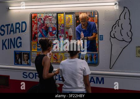 BROOKLYN, NY - 24. JUNI 2026: Senior-Straßenhändler in blauem Hemd reicht Eiscreme-Softeis mit Streuseln an Kunden aus dem Fenster des Trucks Stockfoto