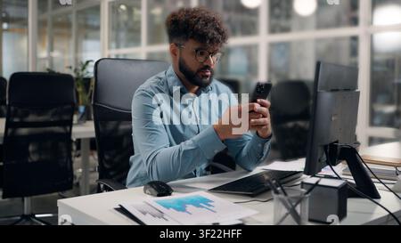Ernster arabischer Geschäftsmann, der auf dem Handy scrollt, Haare berührt, Frisur korrigiert, Locken betrachtet, in die Webcam des Smartphones am Bürotisch schaut Stockfoto