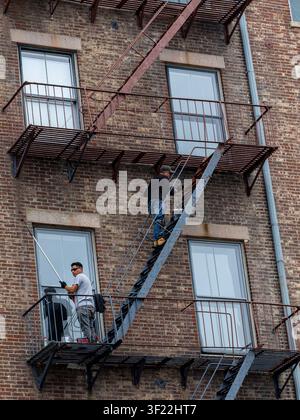 BROOKLYN, NY - 26. JUNI 2026: Zwei Handwerker führen Außenwartungsarbeiten an der eisernen Feuerleiter eines Backsteinwohnhauses durch Stockfoto