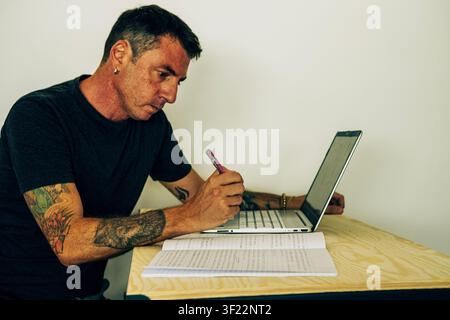 Ein Mann studiert vor einem Computer und einem Notizbuch, während er an einem Schreibtisch sitzt. Der Mann ist zweiundvierzig Jahre alt und trägt ein dunkles T-Shirt Stockfoto