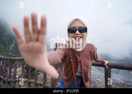 Fröhliche asiatische indonesische Frau mit Hijab und Sonnenbrille greift in Richtung der Kamera, während sie auf einem nebligen Bergausblick steht. Stockfoto