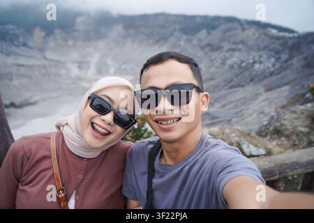 Lächelndes asiatisches Paar mit Sonnenbrille, das ein fröhliches Selfie mit einem malerischen Vulkankrater im Hintergrund macht. Stockfoto