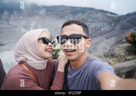 Lächelndes asiatisches Paar mit Sonnenbrille, das ein fröhliches Selfie mit einem malerischen Vulkankrater im Hintergrund macht. Stockfoto