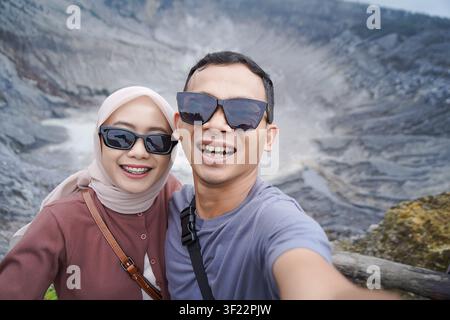 Lächelndes asiatisches Paar mit Sonnenbrille, das ein fröhliches Selfie mit einem malerischen Vulkankrater im Hintergrund macht. Stockfoto