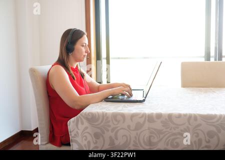 Frau tippt auf einem Laptop, trägt ein Headset, arbeitet aus der Ferne in einer hellen Heimumgebung Stockfoto