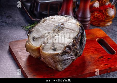 Geräuchertes Störfilet auf einer reichen roten Holzplatte. Dunkle verkohlte Haut kontrastiert mit blassem, flockigem Fleisch. Rosmarin-Zweig-Gewürzgläser im Hintergrund. Stimmungsvolle Lücke Stockfoto
