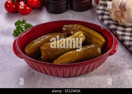 Glänzende grüne Gurken in einer strukturierten roten Schüssel auf grauer Oberfläche. Hintergrund enthält Kirschtomaten, Knoblauchknolle, kariertes Serviette, Gewürzgläser. Warmes Licht Stockfoto