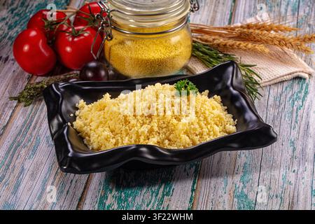 Couscous serviert in schwarzer Keramikschale mit Petersiliengarnitur. Frische Tomaten, Rosmarin, Weizenhalme, Glaskrug mit rohem Couscous darum angeordnet. Rustikal Stockfoto