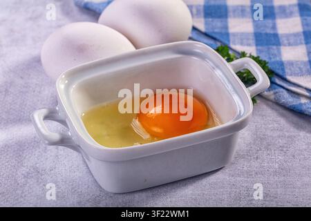 Rohes Eigelb liegt in einer weißen Keramikschale mit ganzen Eiern in der Nähe. Blaues kariertes Tuch und grüne Kräuter setzen Kontraste. Sanfte Beleuchtung betont das leuchtende Orange Stockfoto