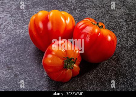 Lebendige rote Heiratomaten mit glänzender Haut, angeordnet auf dunklem grauem Steinboden. Sanfte Studiolichtbeleuchtung betont natürliche Kurven und satte Farbe Stockfoto