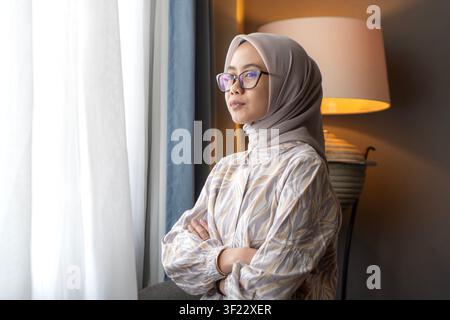 Porträt einer asiatischen indonesischen muslimischen Frau mit Hijab und Brille, die lächelnd aus einem hellen Fenster im Innenraum blickt. Stockfoto