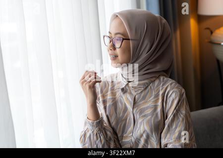Porträt einer asiatischen indonesischen muslimischen Frau mit Hijab und Brille, die lächelnd aus einem hellen Fenster im Innenraum blickt. Stockfoto