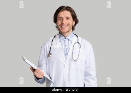 Lächelnder junger Arzt im weißen Mantel mit Stethoskop und Klemmbrett Stockfoto