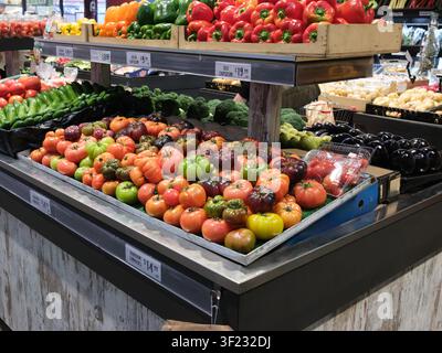 Eine üppige Auswahl an farbenfrohen Heirloom-Tomaten liegt unter Regalen, die mit frischen Paprikaschoten gestapelt sind, in einem Supermarkt. Stockfoto