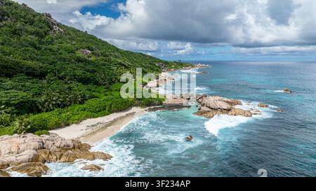Wellen schlagen gegen die felsige Küste, umgeben von üppiger tropischer Vegetation und türkisfarbenem Meereswasser. La Digue, Seychellen. Stockfoto