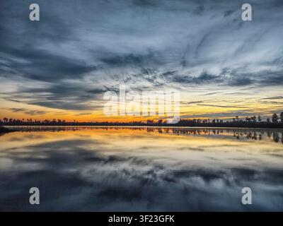 Dramatischer Himmel spiegelt sich in ruhigem Wasser. Goldenes Licht bahnt sich seinen Weg durch dunkle Wolken. Silhouetten von Bäumen säumen die entfernte Uferlinie. Die stille Oberfläche spiegelt den Himmel wider v Stockfoto