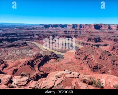 Ein weites Canyon erstreckt sich unter klaren blauen Himmeln. Ein Fluss windet sich durch uralte rote Felsformationen. Sonnenlicht betont schroffe Klippen und tiefe Täler Stockfoto