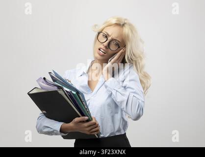 Gestresste junge kaukasische Frau, die Ordner hält und sich im Büro das Gesicht berührt, Geschäftskonzept Stockfoto