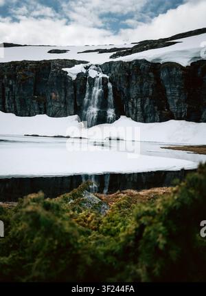 Wasserfall in schneebedeckten Bergen mit gefrorener Seelandschaft in Norwegen, stimmungsvolle Landschaft in Skandinavien, Frühlingsklima, Schmelzeis, malerische Natur, Reiseszenerie Stockfoto