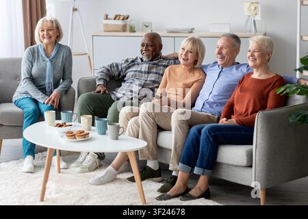 Fröhliche diverse Seniorengruppe genießt gemeinsam eine Fernsehsendung im Wohnzimmer, entspanntes Wochenende Stockfoto