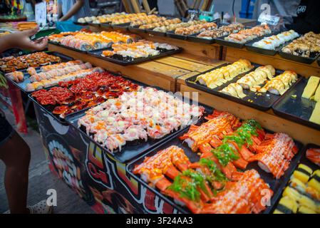 Eine lebhafte Auslage mit verschiedenen Sushi-Sorten ist auf Tabletts an einem geschäftigen Marktstand ausgelegt und präsentiert farbenfrohe Rolls, Sashimi und Meeresfrüchte-Delikateszen für den Laden Stockfoto