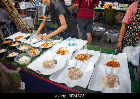 Eine geschäftige Nachtmarkt-Szene mit einem Stand, auf dem eine Vielzahl traditioneller koreanischer Gerichte auf weißen Pappschalen arrangiert ist, während Menschen zubereiten und s Stockfoto