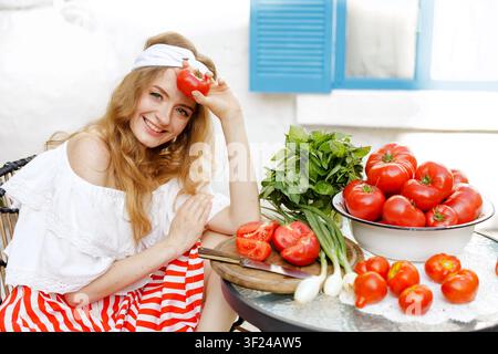 Eine fröhliche Frau hält eine reife Tomate, umgeben von frischen Lebensmitteln, bereit für die Zubereitung von Gazpacho und Salsa, und zeigt die lebendige Erntesaison. Stockfoto