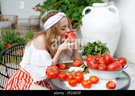 Eine Frau genießt eine frische reife Tomate, umgeben von einer Tomaten- und Basilharveste, perfekt für die Zubereitung von Gazpacho, Salsa oder Tomatensoße. Stockfoto