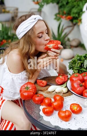 Eine Frau genießt eine reife Tomate, umgeben von frischen Zutaten für Salsa, Gazpacho und Tomatensuppe in einer lebendigen Gartenumgebung. Stockfoto