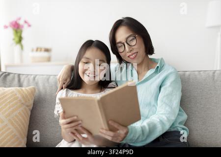Zufriedene asiatische Mutter liest ein Buch mit ihrer Tochter zu Hause, lächeln gemeinsam, Familienkonzept Stockfoto