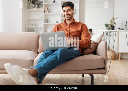 Lächelnder junger Mann mit Kopfhörern, der zu Hause am Laptop arbeitet, Geschäftskonzept Stockfoto