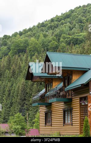 Ein mehrstöckiges Holzblockhaus mit Eisenbalkonen steht an einem steilen Hang, der mit dichtem Nadel- und Laubwald bedeckt ist. Stockfoto