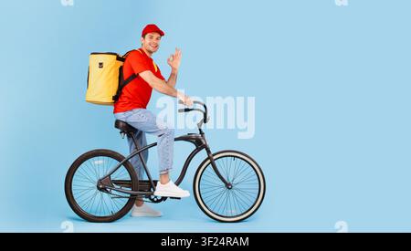 Lächelnder männlicher Lieferfahrer in rotem Hemd fährt Fahrrad, städtische Umgebung, Konzept für Lebensmittellieferung Stockfoto