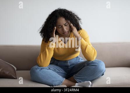 Junge schwarze Frau mit Kopfschmerzen, die auf der Couch sitzt, häusliche Umgebung, Stresskonzept Stockfoto