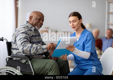 Lächelnder älterer afroamerikanischer Mann im Rollstuhl, der mit einer Krankenschwester in der Klinik spricht, Gesundheitskonzept Stockfoto