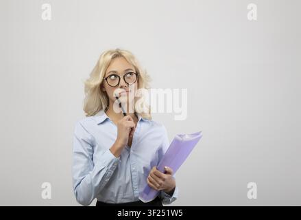 Nachdenkliche junge Frau, die Ordner und Stift in einer Büroumgebung hält, Geschäftskonzept Stockfoto