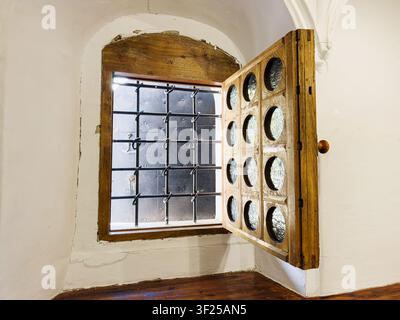 Rustisches Holzfenster mit Metallgitter, alte Innentür mit Lüftungsschlitzen, gewölbte Decke, polierter Holzboden, hellbeige Wände, dunkelbraun Stockfoto