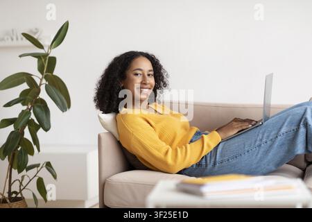 Lächelnde schwarze Frau arbeitet an einem Laptop, entspannt sich auf der Couch zu Hause, gemütlicher Lebensstil Stockfoto
