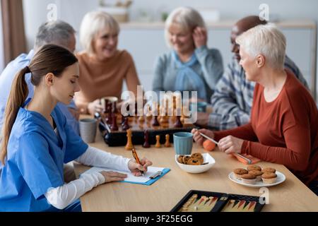 Konzentrierte junge kaukasische Frau macht Notizen, während sie in einer Senioreneinrichtung Schach spielt Stockfoto