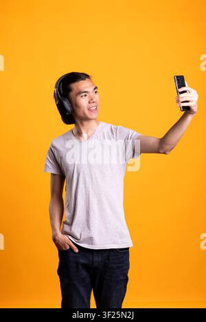 Asiatischer Mann mit Kopfhörern steht im Studio mit gelbem Hintergrund und vloggt tägliche Aktivitäten. Selbstbewusster männlicher Model hebt die Hand, während er ein Selfie macht, lächelt und posiert lässig für das Publikum. Stockfoto