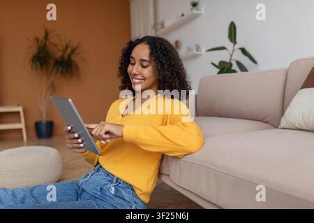 Lächelnde junge afroamerikanische Frau, die ein Tablet benutzt, während sie zu Hause auf der Couch sitzt Stockfoto