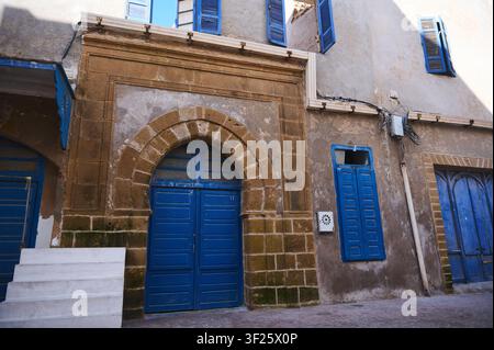 Essaouira, Marrakesch - Safi-Region, Marokko, 10. Juli 2026: Bunte blaue Türen und Fensterläden in verwitterten Steinbögen entlang einer engen Gasse Stockfoto