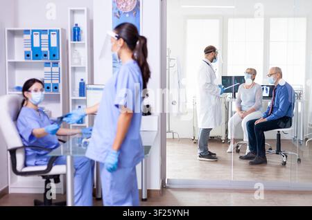 Medizinisches Personal trägt Gesichtsmasken und arbeitet in einem Krankenhausflur, während ein Arzt in einem Untersuchungsraum mit einem alten Ehepaar über die Pflege spricht. Stockfoto